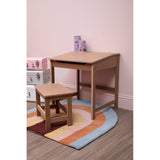 Natural wood desk and matching stool set for children, positioned on a colorful, circular rug. The desk features a simple, rectangular design with clean lines, while the stool is smaller and complements the desk's height. The background includes a soft pink wall, enhancing the playful atmosphere