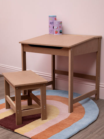 Natural wood desk and matching stool set designed for children, featuring a simple and sturdy construction. The desk has a smooth surface and a small drawer, while the stool is low to the ground for easy access. Both pieces are placed on a colorful, circular rug with a pastel design. In the background, there are a few small, colorful blocks stacked on the desk