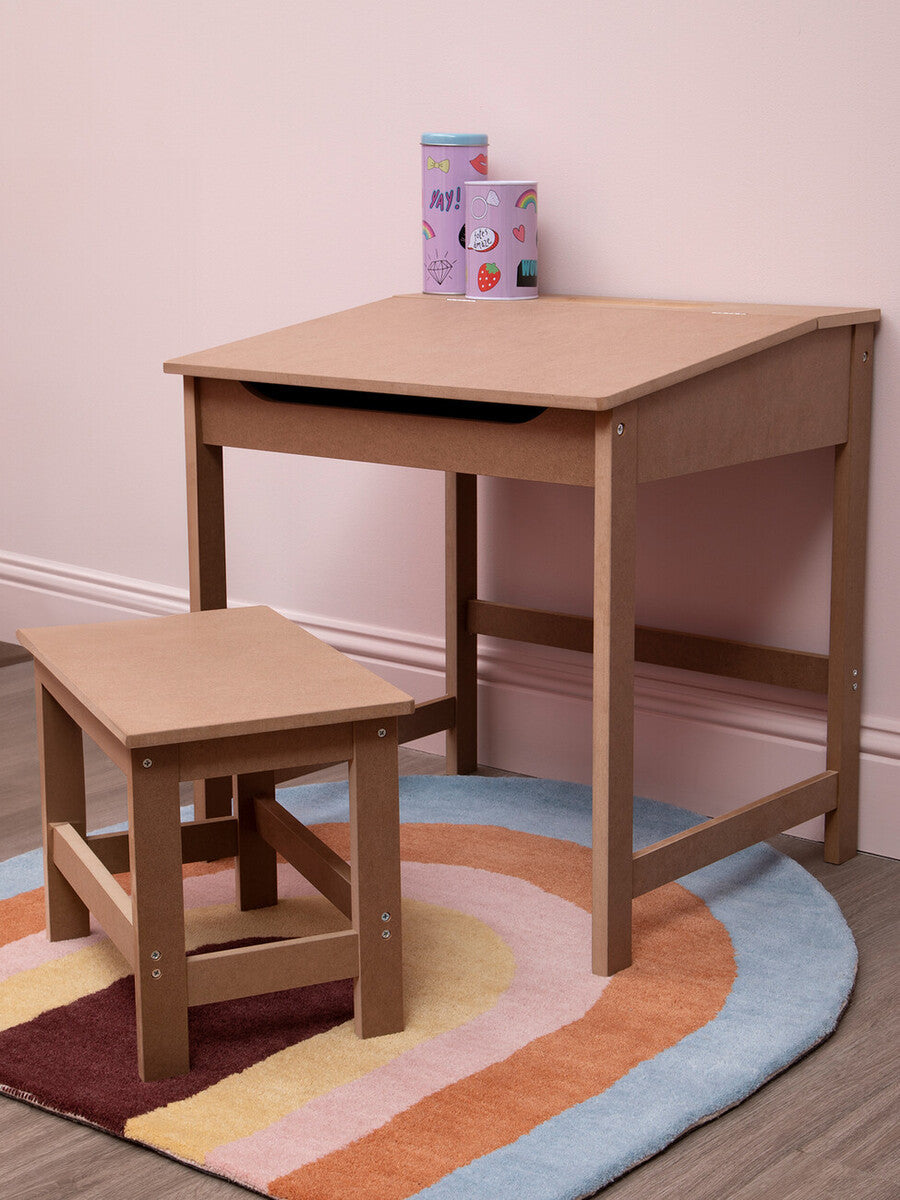 Natural wood desk and matching stool set designed for children, featuring a simple and sturdy construction. The desk has a smooth surface and a small drawer, while the stool is low to the ground for easy access. Both pieces are placed on a colorful, circular rug with a pastel design. In the background, there are a few small, colorful blocks stacked on the desk