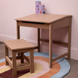 Natural wood desk and matching stool set designed for children, featuring a simple, sturdy construction. The desk has a single drawer and is placed on a colorful circular rug, with two small books stacked on top. The wall behind is painted a soft color, creating a cozy atmosphere