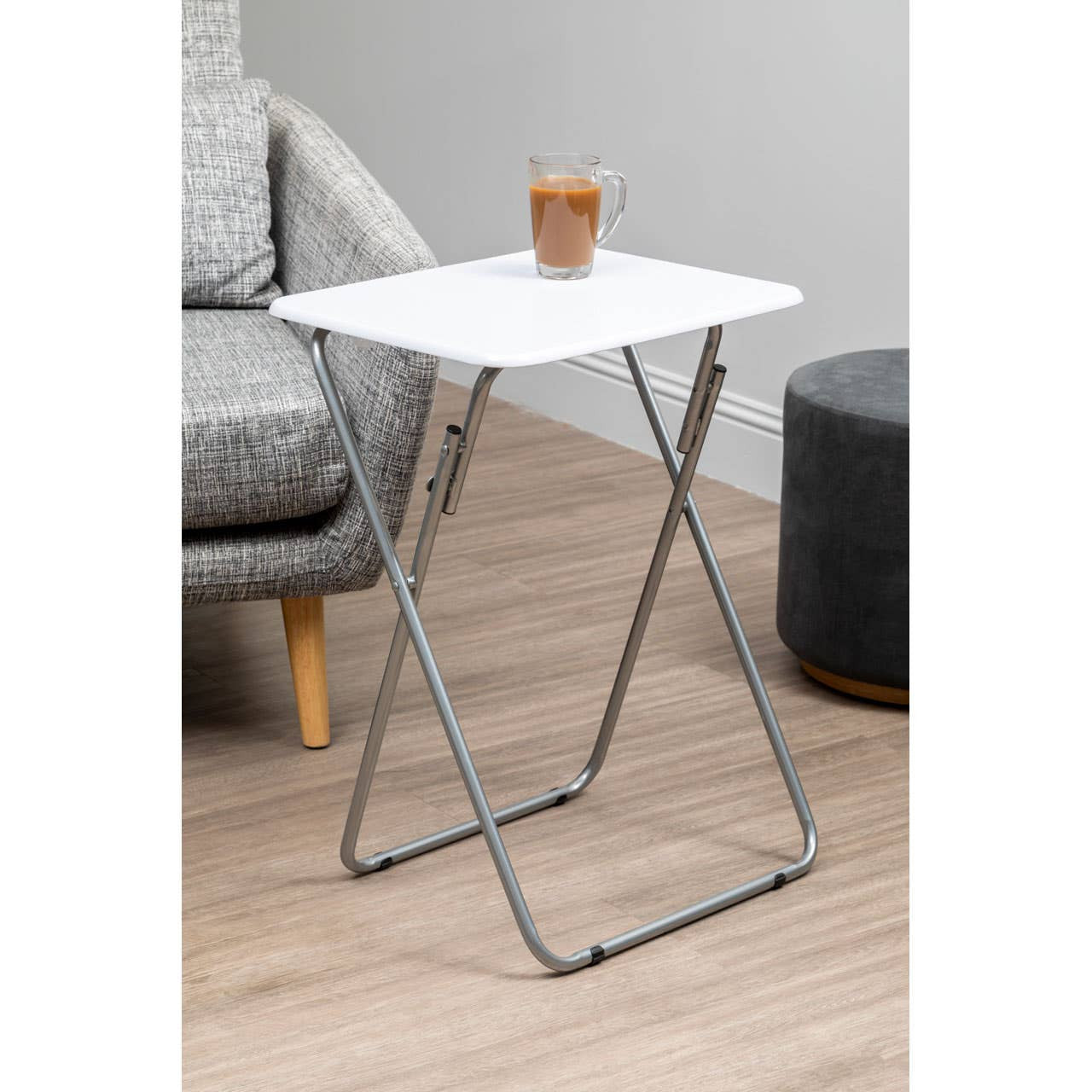 A folding table with a white top and silver metal legs, positioned next to a gray sofa and a dark round side table, with a small plant in a brown pot placed on the table's surface. The floor is light wood