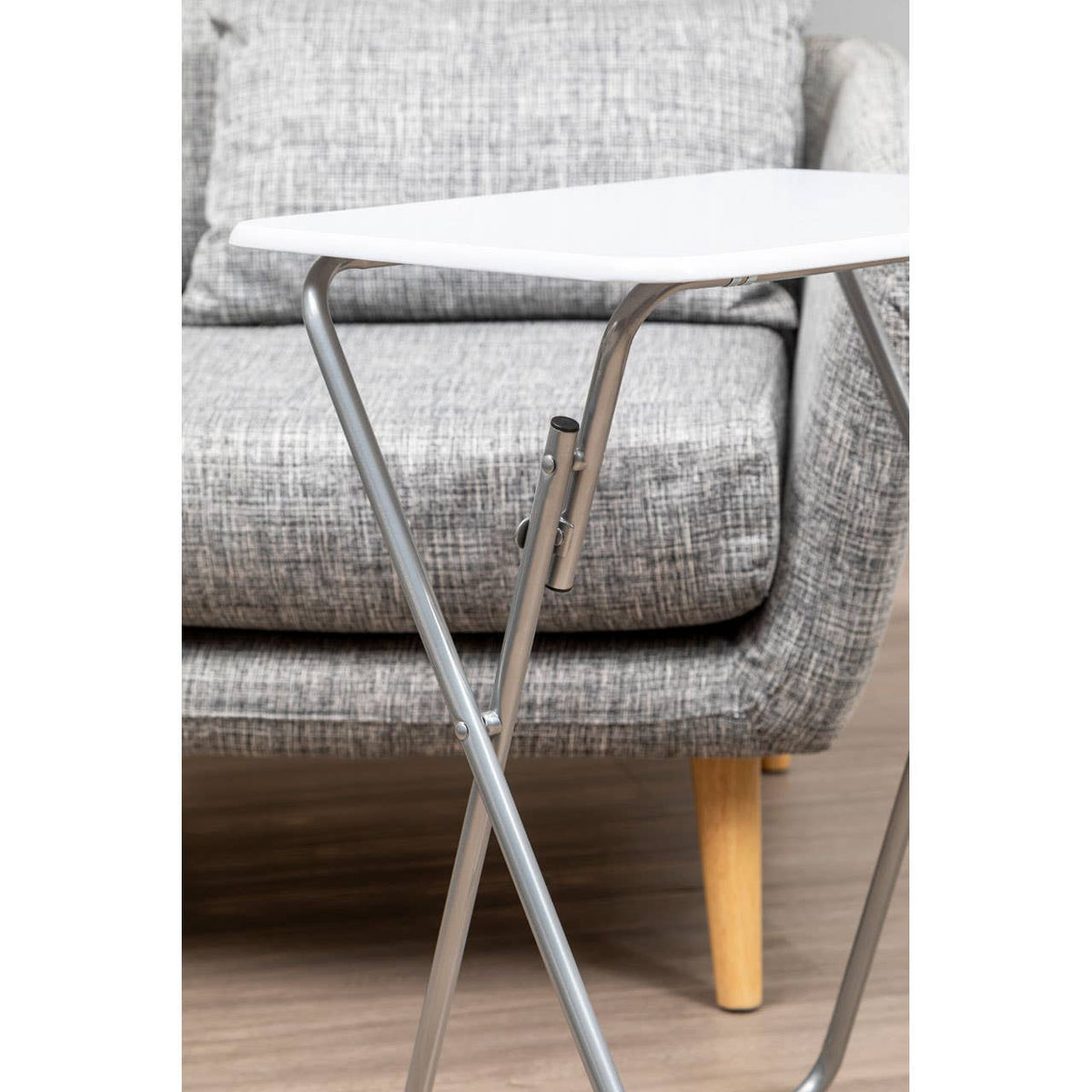 Folding table with a white top and silver metal legs positioned next to a gray upholstered sofa with wooden legs. The table is compact and designed for easy storage, complementing the modern living room setting