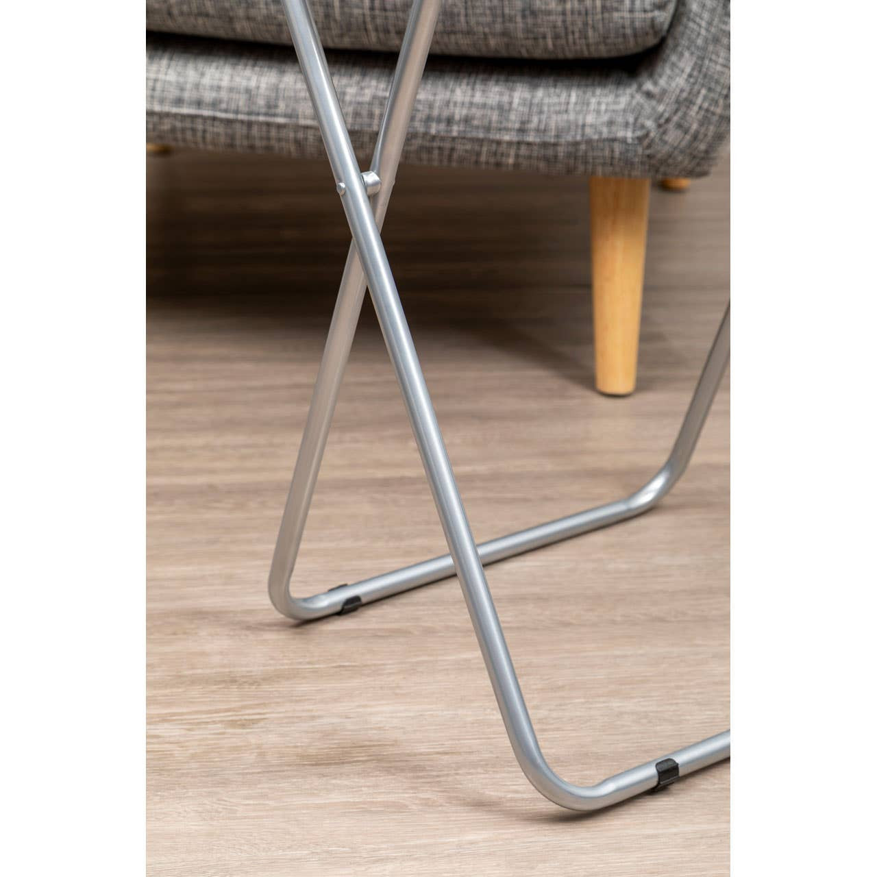 Folding table with a white top and silver metal legs, positioned on a wooden floor, partially visible next to a textured gray sofa