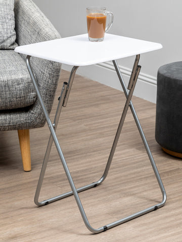 Folding table with a white top and silver metal legs, positioned next to a gray upholstered sofa, featuring a glass of beverage on top, with a dark round side table visible in the background on a light wooden floor
