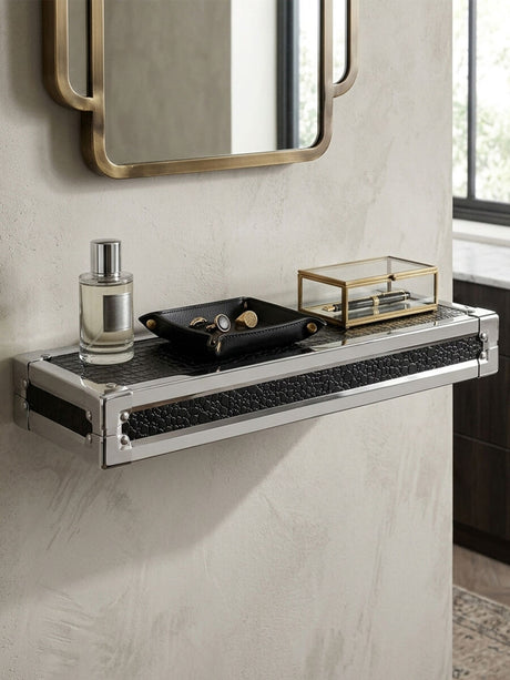 Small wall shelf with a crocodile leather effect finish, mounted on a light-colored wall. The shelf holds a decorative black dish, a glass container with a gold lid, and a bottle of perfume. A round mirror with a gold frame is visible above the shelf, reflecting the room's interior