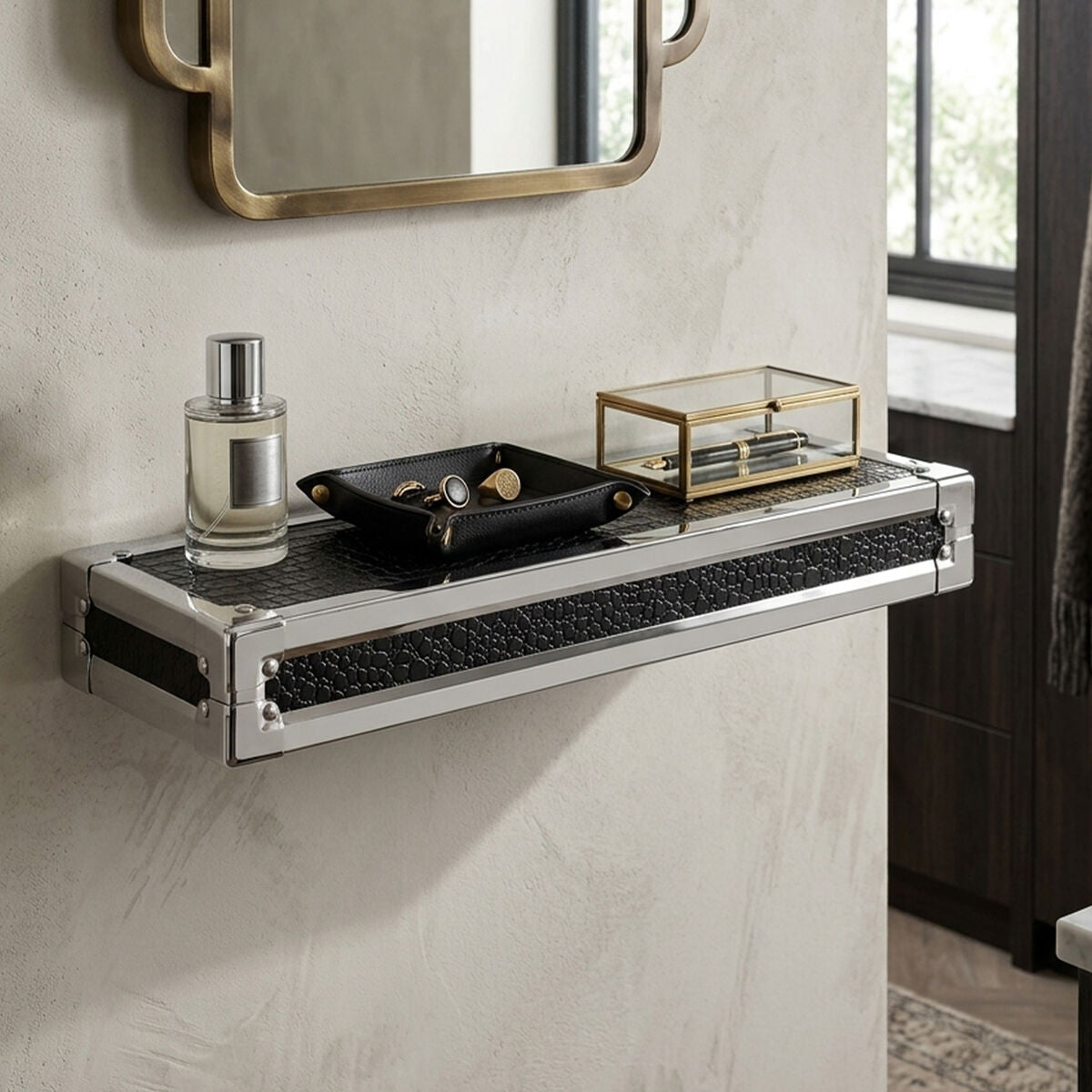 Small wall shelf featuring a crocodile leather effect, mounted against a textured wall. The shelf holds a decorative black tray with small items, a glass box, and a bottle of perfume. A round mirror with a gold frame is visible above the shelf, reflecting light from a nearby window