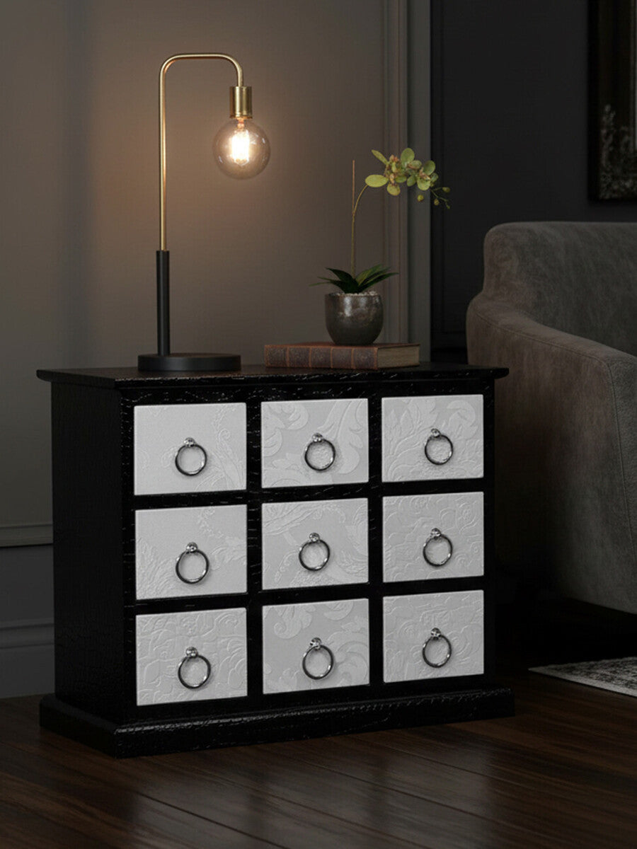 Rectangular cabinet featuring nine drawers with a crocodile leather effect, set against a dark background. The cabinet has a sleek black frame and white drawer fronts, each adorned with a circular handle. A warm light bulb hangs above, illuminating the cabinet, while a small potted plant adds a touch of greenery to the scene. The cabinet is positioned next to a sofa, enhancing the cozy atmosphere of the room