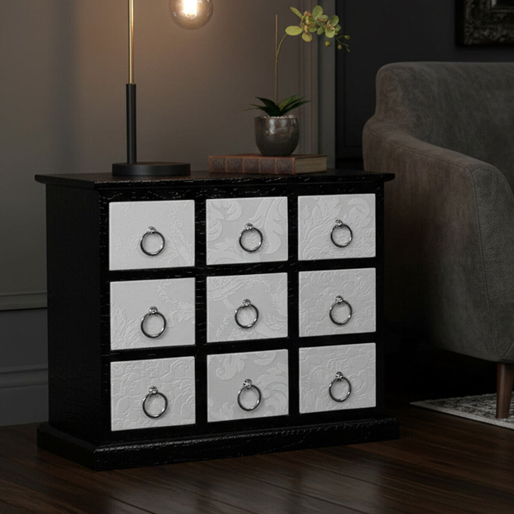 A rectangular cabinet featuring nine drawers with a crocodile leather effect design, set against a dark background. The cabinet has a sleek black frame and white drawer fronts, each adorned with a round metal handle. A candle is lit on top, casting a warm glow, while a small plant adds a touch of greenery beside the cabinet. The floor is wooden, complementing the overall aesthetic