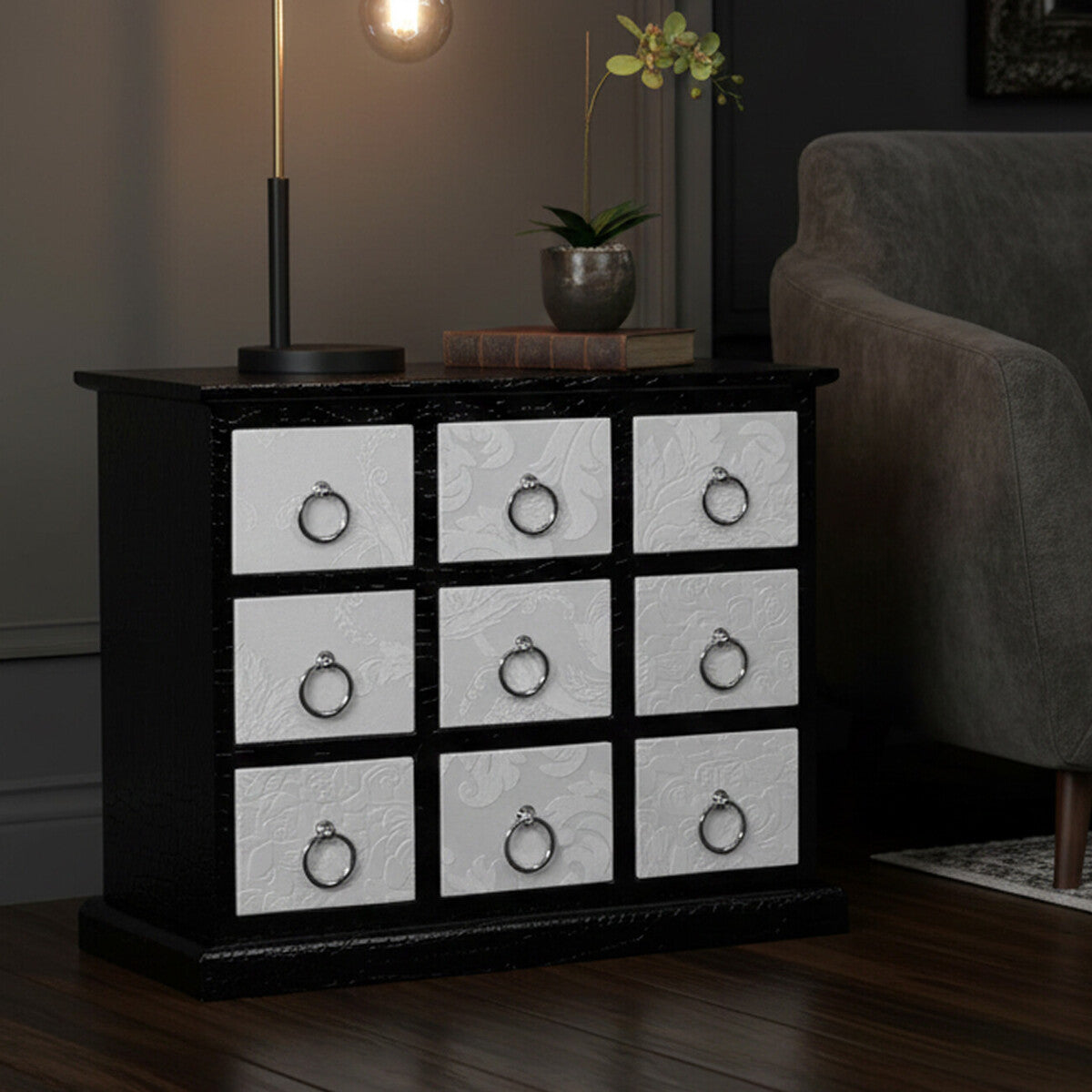 A rectangular cabinet featuring nine drawers with a crocodile leather effect design, set against a dark background. The cabinet has a sleek black frame and white drawer fronts, each adorned with a round metal handle. A candle is lit on top, casting a warm glow, while a small plant adds a touch of greenery beside the cabinet. The floor is wooden, complementing the overall aesthetic