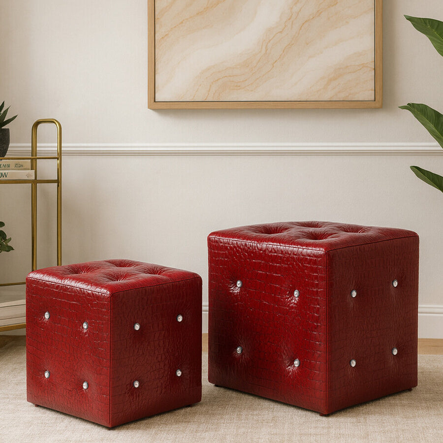 Two red stools with a crocodile leather effect, one larger and one smaller, placed on a light-colored floor. The stools feature decorative silver studs on their surfaces, and a neutral-toned wall art piece is visible in the background, along with a gold metal side table and a green plant