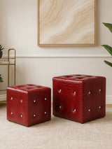 Two red stools with a crocodile leather effect, one larger and one smaller, placed on a light-colored floor. The stools feature decorative silver studs on their surfaces, and a neutral-toned wall art piece is visible in the background, along with a gold metal side table and a green plant