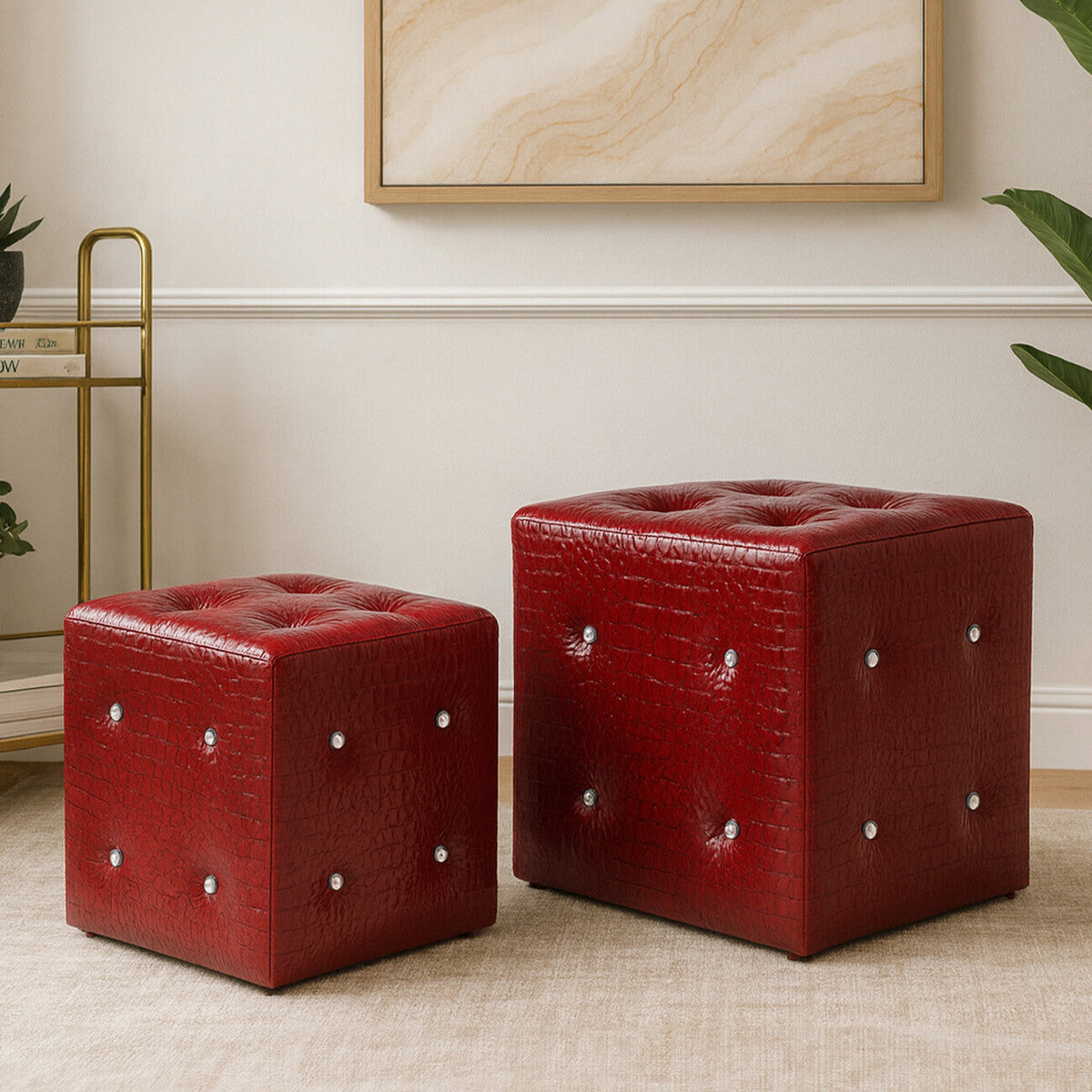 Two red stools with a crocodile leather effect, one larger and one smaller, featuring tufted tops and decorative silver studs. They are placed on a light-colored floor against a neutral wall, with a piece of abstract art visible in the background