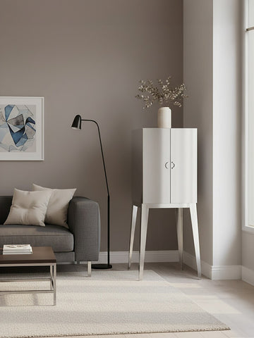 A sleek stainless steel two-door cabinet stands in a modern living room, featuring a minimalist design with clean lines. The cabinet is positioned next to a gray sofa adorned with decorative pillows, and a floor lamp casts soft light nearby. A piece of abstract artwork hangs on the wall, complementing the neutral color palette of the room, which includes a light-colored rug and large windows allowing natural light to fill the