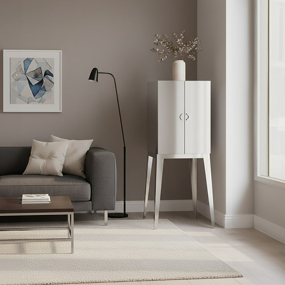 A modern living room featuring the Lure Stainless Steel 2 Door Cabinet with a sleek white finish and minimalist design, positioned beside a gray sofa adorned with decorative pillows. A floor lamp stands nearby, and a piece of artwork hangs on the wall, complementing the neutral color palette of the space. Natural light streams in through large windows, illuminating the room