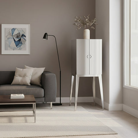 A modern living room featuring the Lure Stainless Steel 2 Door Cabinet with a sleek white finish and minimalist design, positioned beside a gray sofa adorned with decorative pillows. A floor lamp stands nearby, and a piece of artwork hangs on the wall, complementing the neutral color palette of the space. Natural light streams in through large windows, illuminating the room