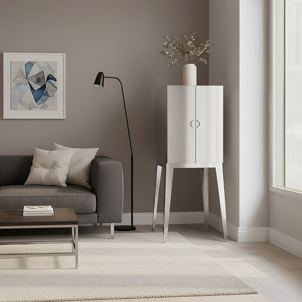 A modern living room featuring the Lure Stainless Steel 2 Door Cabinet with a sleek white finish and minimalist design, positioned beside a gray sofa adorned with decorative pillows. A floor lamp stands nearby, and a piece of artwork hangs on the wall, complementing the neutral color palette of the space. Natural light streams in through large windows, illuminating the room