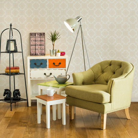 A black three-tier shelf unit stands against a light-colored wall, showcasing various decorative items. Next to it, a green upholstered armchair with button detailing is positioned beside a small white side table. The side table holds a potted plant and a decorative bowl. A floor lamp with a white shade illuminates the space, while a colorful storage unit with orange and blue drawers adds a pop of color to