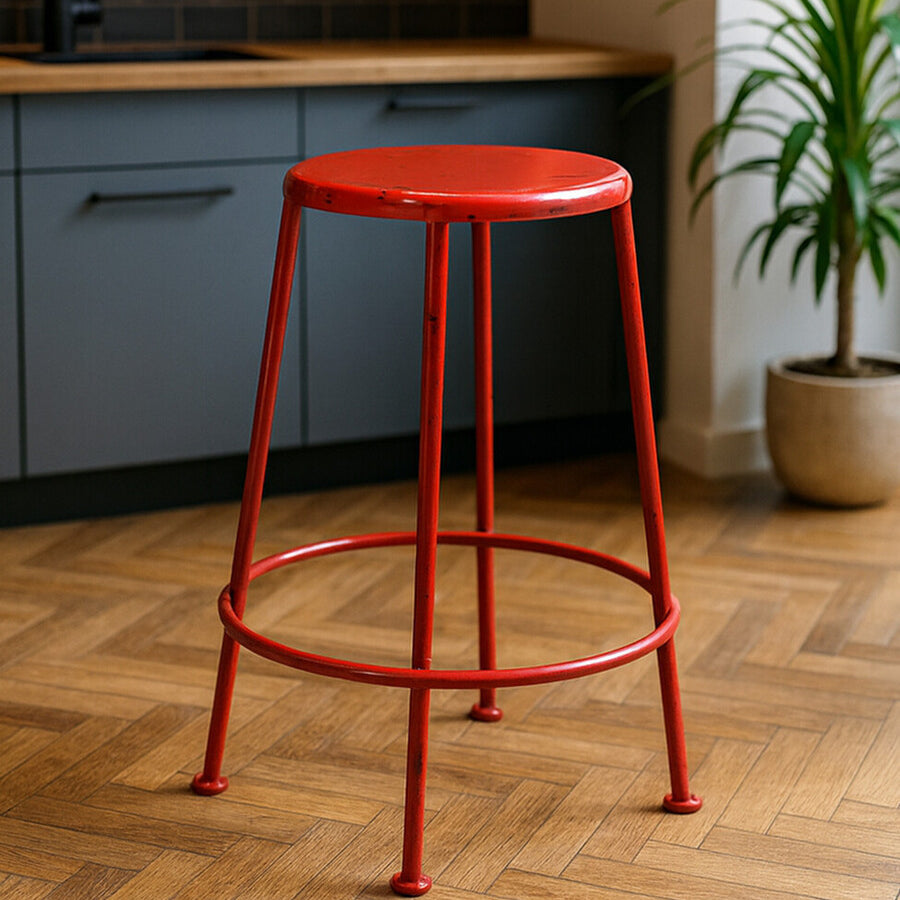 A bright red metal stool with a round seat and slender legs, positioned on a wooden floor in a modern kitchen setting with gray cabinetry and a potted plant in the background