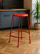 A bright red metal stool with a round seat and slender legs, positioned on a wooden floor in a modern kitchen setting with gray cabinetry and a potted plant in the background