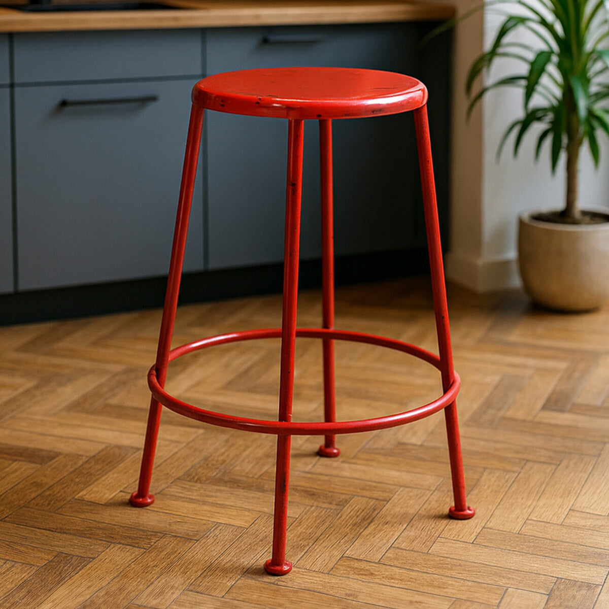 Bright red metal stool with a round seat and four slender legs, set against a wooden floor and a kitchen backdrop featuring gray cabinetry and a potted plant