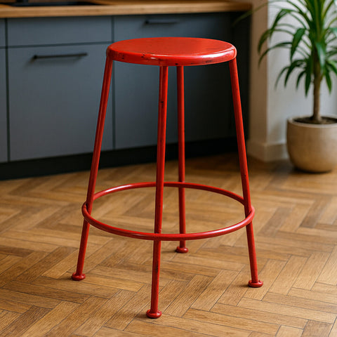 Bright red metal stool with a round seat and four slender legs, set against a wooden floor and a kitchen backdrop featuring gray cabinetry and a potted plant