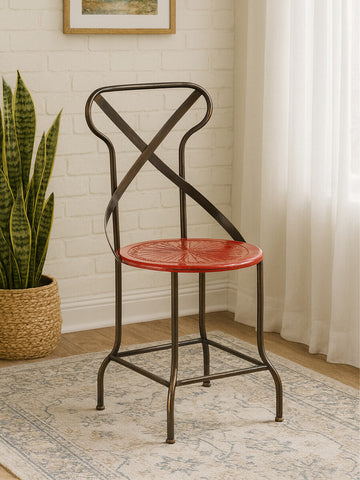 A red metal seat chair with a cross-back design, featuring a round seat and slender legs, positioned on a light-colored rug in a well-lit room with a plant and curtains in the background