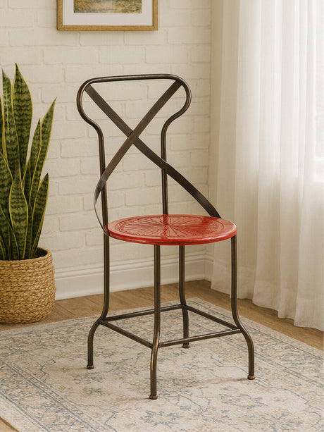 A red metal seat chair with a cross-back design, featuring a round seat and slender legs, positioned on a light-colored rug in a well-lit room with a plant and curtains in the background