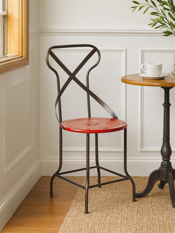 A red metal seat chair with a crisscross backrest, positioned next to a small round wooden table. The chair features a sleek design and is placed on a wooden floor, with natural light coming through a nearby window. A cup is resting on the table, and there are decorative elements in the background