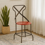 A red metal seat chair with a cross-back design, featuring a round seat and slender black legs, positioned next to a potted plant and a light-colored rug in a bright room with natural light filtering through sheer curtains