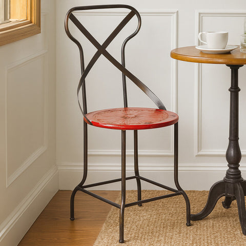 A red metal seat chair with a cross-back design, positioned next to a small wooden side table. The chair features a round seat and slender black legs, set against a light-colored wall and wooden floor