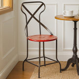 A red metal seat chair with a cross-back design, positioned next to a small wooden side table. The chair features a round seat and slender black legs, set against a light-colored wall and wooden floor