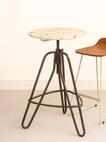 Cream metal bar table with a round top and a unique, geometric base featuring slender legs. The table is positioned on a light-colored floor, with a wooden chair partially visible in the background