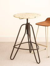Cream metal bar table with a round top and a unique, geometric base featuring slender legs. The table is positioned on a light-colored floor, with a wooden chair partially visible in the background