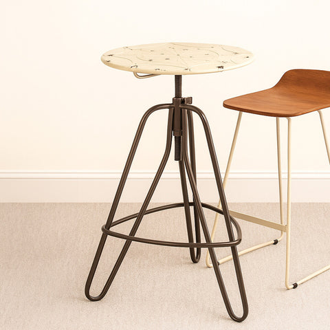 Cream metal bar table with a round top and a unique textured surface, supported by a sleek, dark metal tripod base. The table stands on a light-colored carpet, with a wooden bar stool partially visible in the background