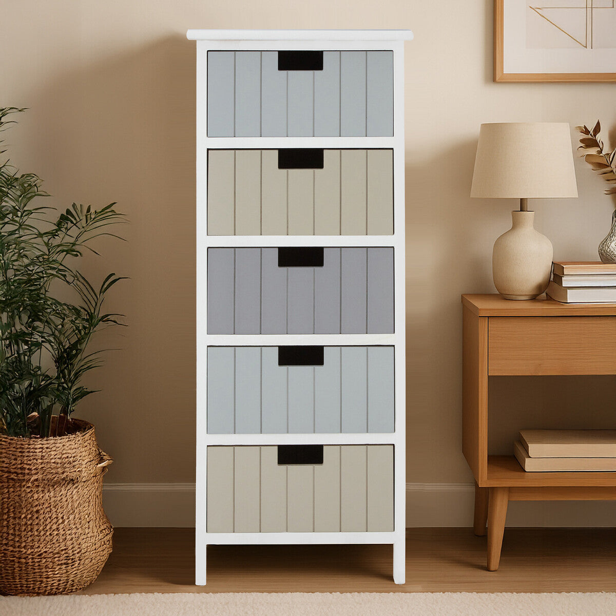 A tall, white chest of drawers featuring five fabric storage bins in varying shades of gray and blue. The chest is positioned next to a wooden side table with a lamp and a decorative plant in a woven basket. The background includes a framed picture on the wall