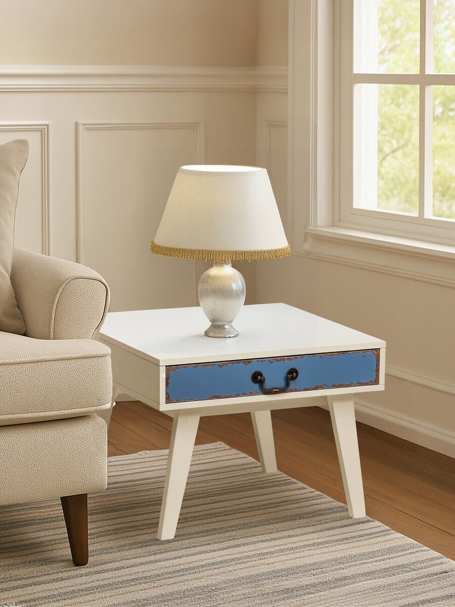 A stylish side table with a white finish and a blue drawer, featuring tapered wooden legs. A lamp with a cream-colored shade sits on top, next to a beige sofa in a well-lit room with light-colored walls and a window