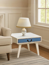 A stylish side table with a white finish and a blue drawer, featuring tapered wooden legs. A lamp with a cream-colored shade sits on top, next to a beige sofa in a well-lit room with light-colored walls and a window