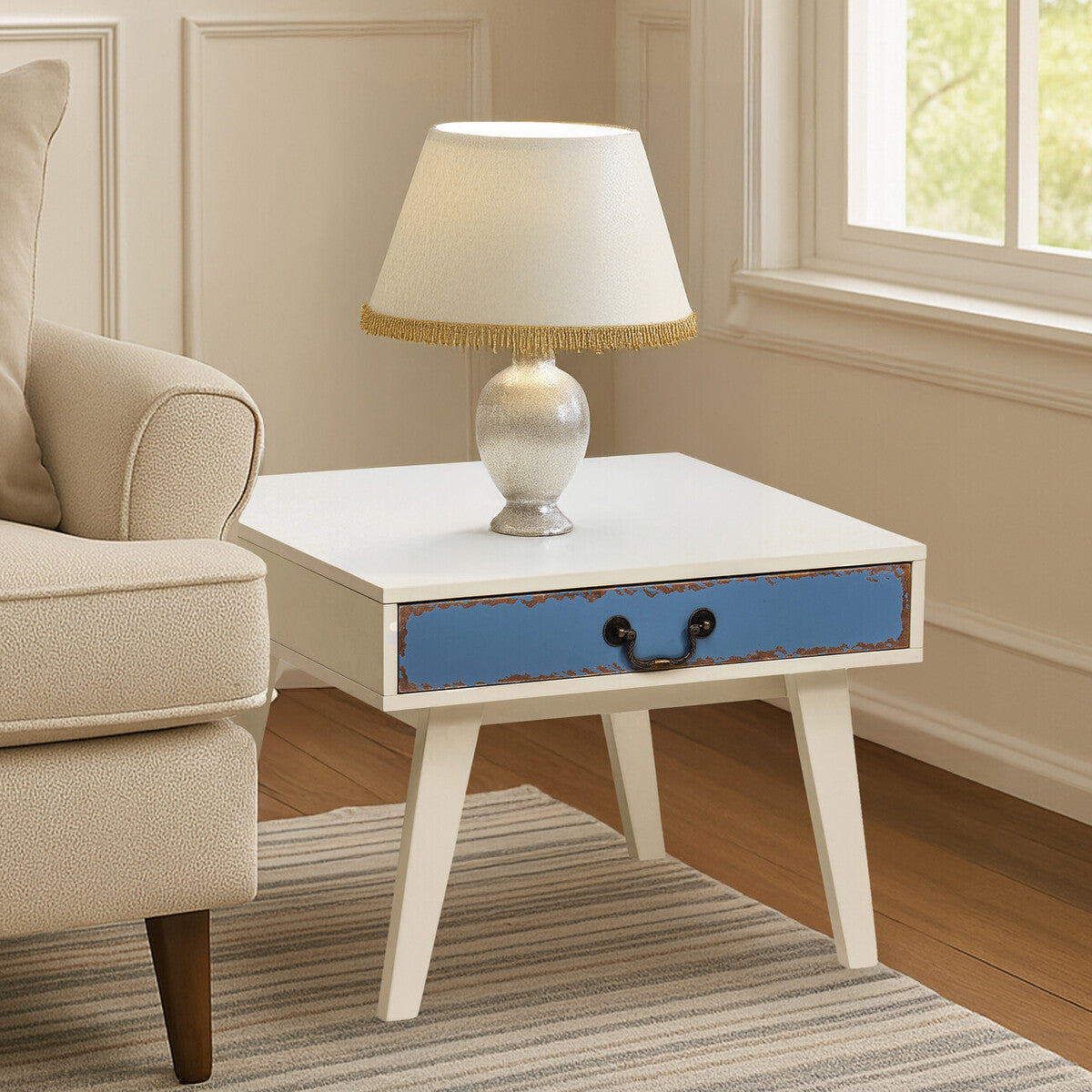 A stylish side table with a white top and a blue drawer, featuring a gold handle. The table has four tapered wooden legs and is positioned next to a light-colored sofa. A lamp with a cream shade and a decorative base sits on top of the table, with a soft rug underneath. Natural light streams in from a nearby window