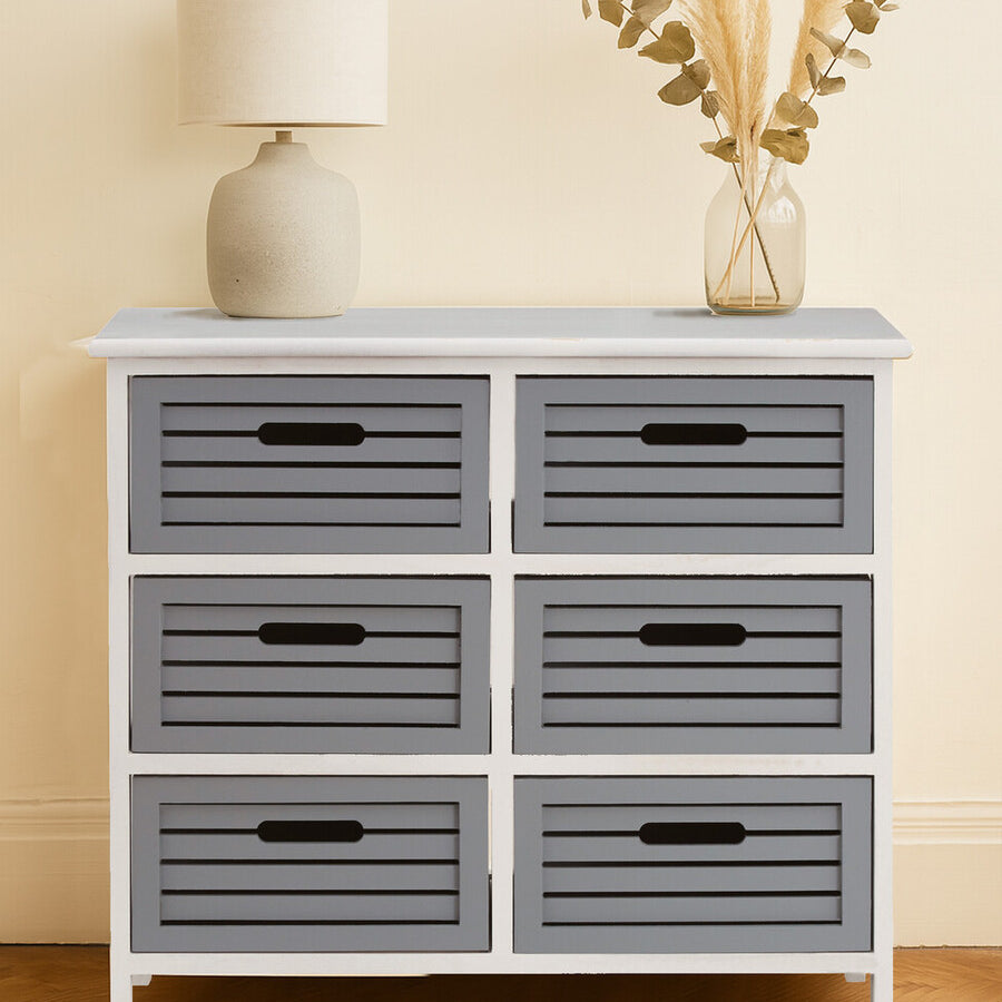 A stylish furniture unit featuring six grey drawers with horizontal slats, set against a light-colored wall. The unit has a white frame and black handles on each drawer. A lamp and a decorative arrangement of dried plants are placed on top, adding a cozy touch to the overall design. The floor is wooden, complementing the furniture's aesthetic