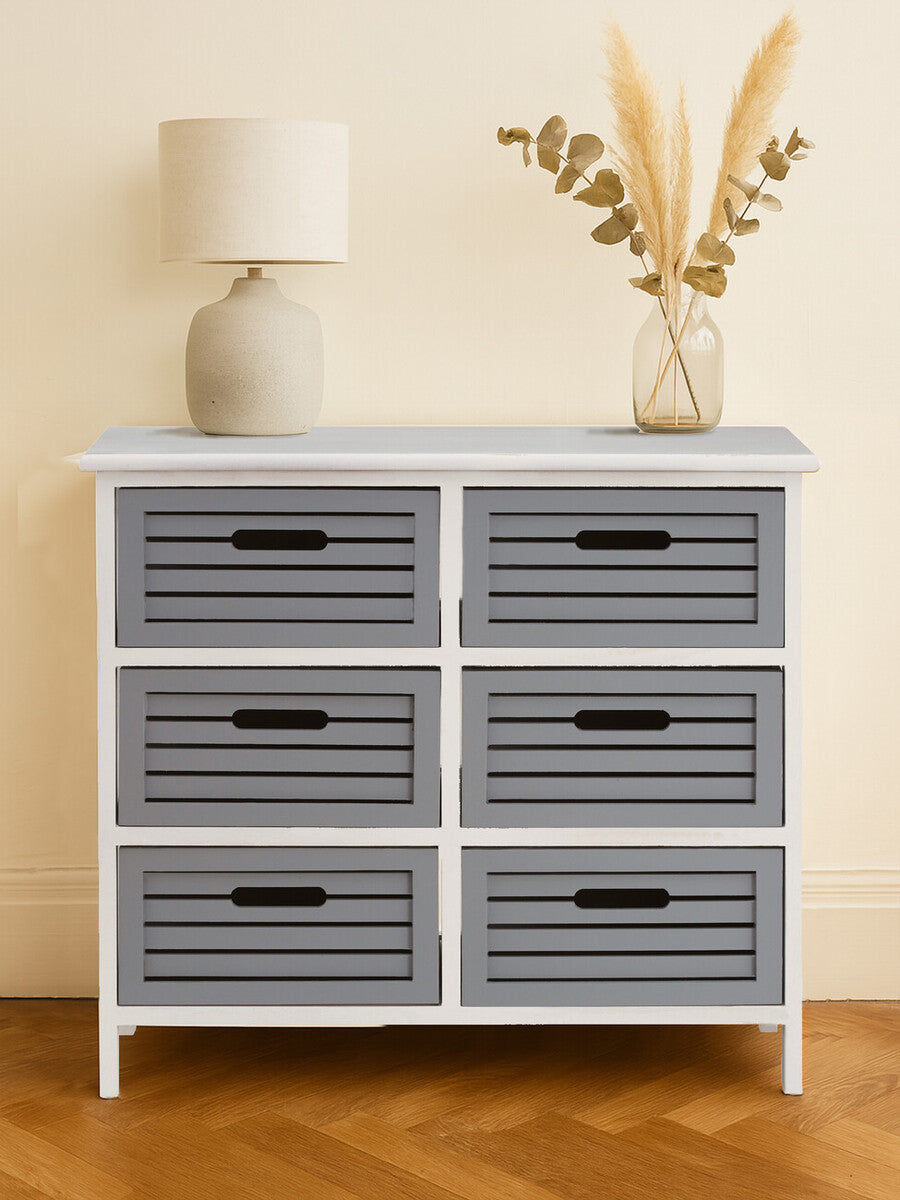 A stylish furniture unit featuring six grey drawers with horizontal slats, set against a light-colored wall. The unit has a white frame and black handles on each drawer. A lamp and a decorative arrangement of dried plants are placed on top, adding a cozy touch to the overall design. The floor is wooden, complementing the furniture's aesthetic
