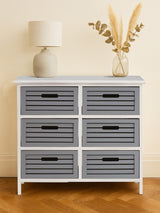A stylish furniture unit featuring six grey drawers with horizontal slats, set against a light-colored wall. The unit has a white frame and black handles on each drawer. A lamp and a decorative arrangement of dried plants are placed on top, adding a cozy touch to the overall design. The floor is wooden, complementing the furniture's aesthetic