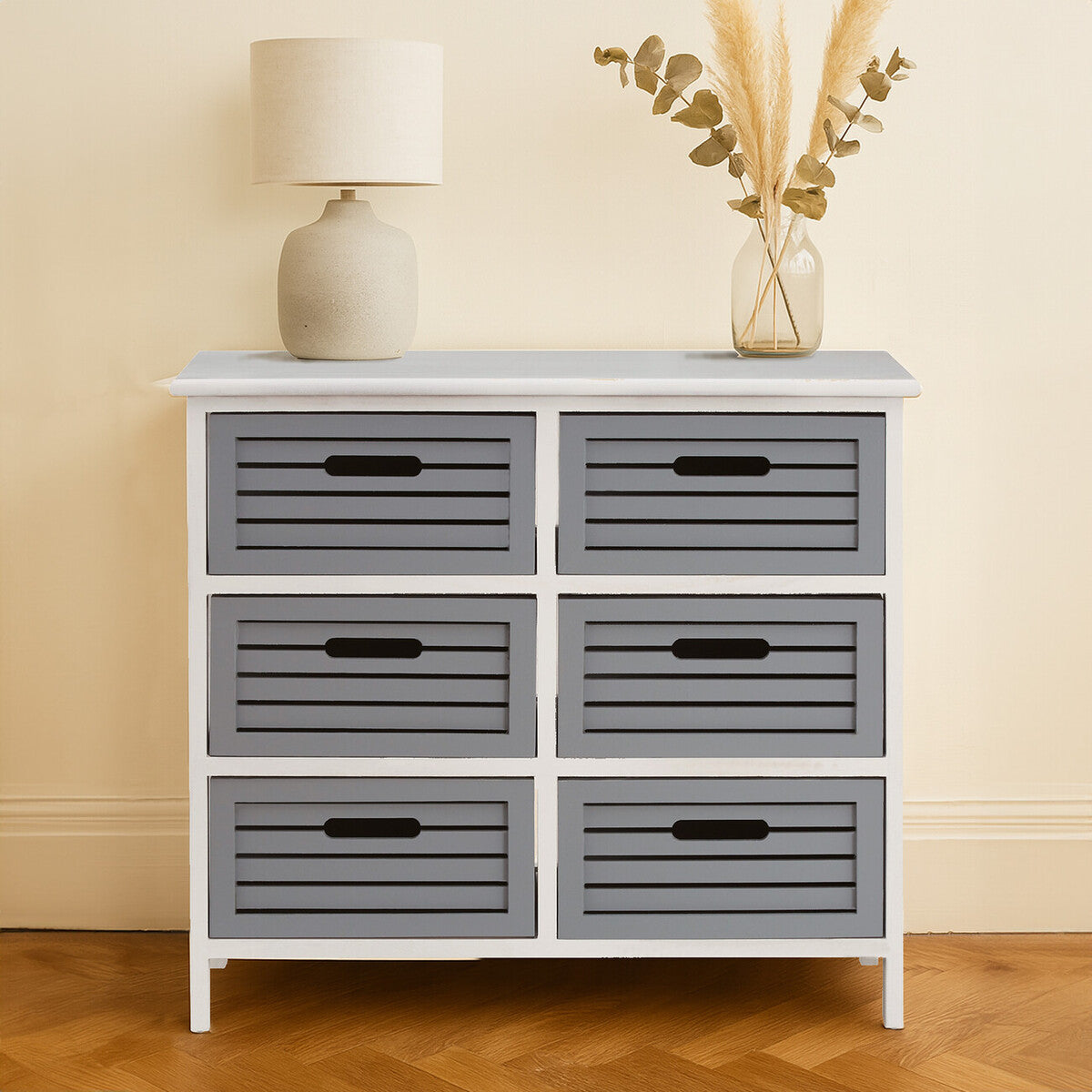 A white furniture unit with six grey drawers, featuring horizontal slats and black handles, placed against a light-colored wall. A beige lamp and a decorative vase with dried flowers are positioned on top of the unit, with a wooden floor visible beneath