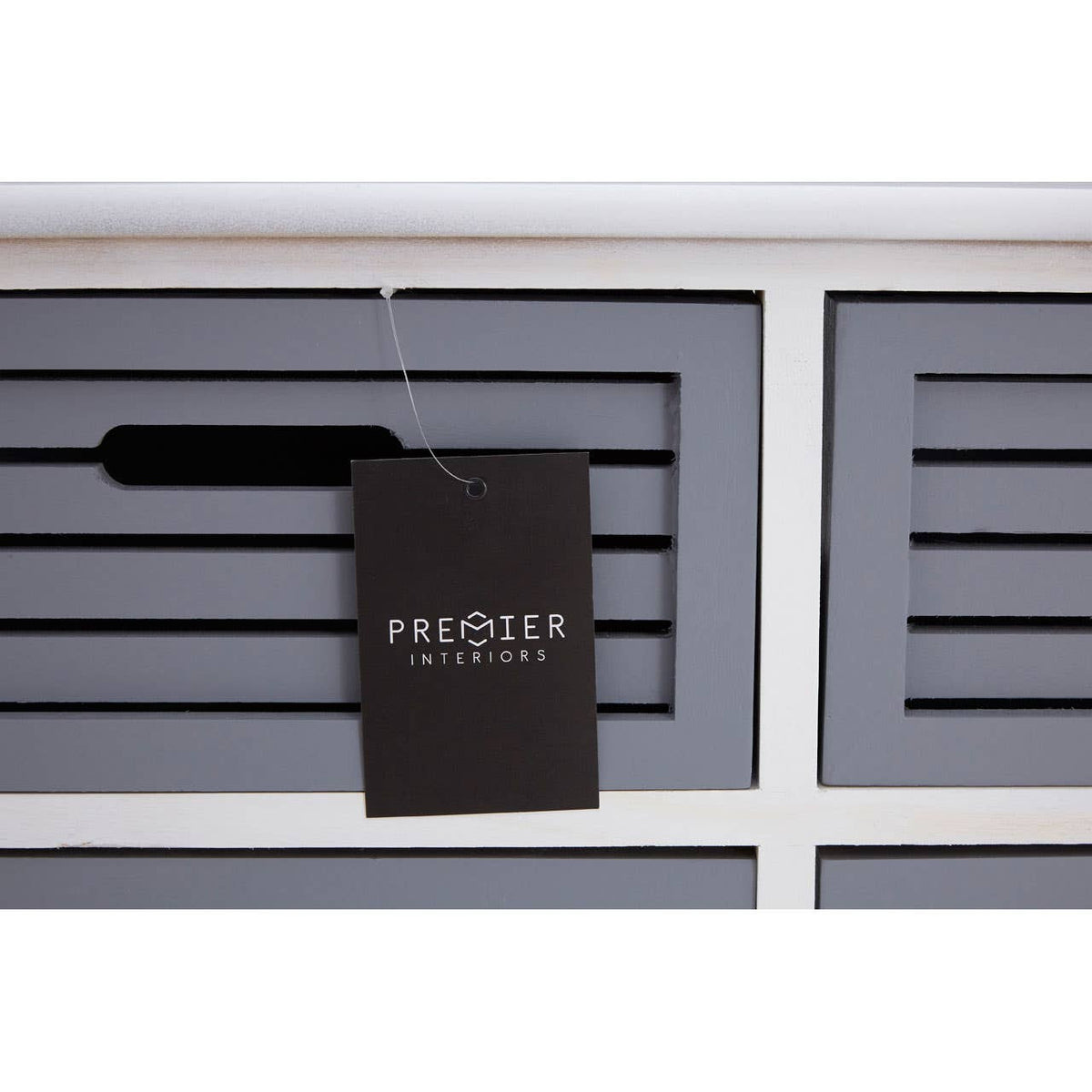 A close-up view of a grey drawer unit featuring six horizontal drawers with slatted fronts. A black tag hangs from one of the drawers, displaying the brand name. The unit has a clean, modern design with a white top and a smooth finish