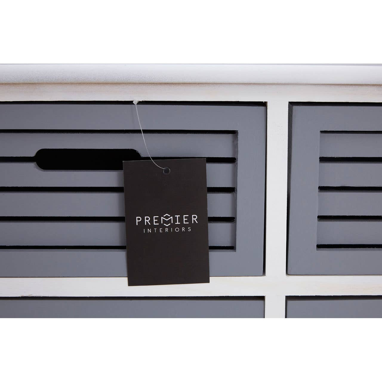 A close-up view of a grey drawer unit featuring six horizontal drawers with slatted fronts. A black tag hangs from one of the drawers, displaying the brand name. The unit has a clean, modern design with a white top and a smooth finish