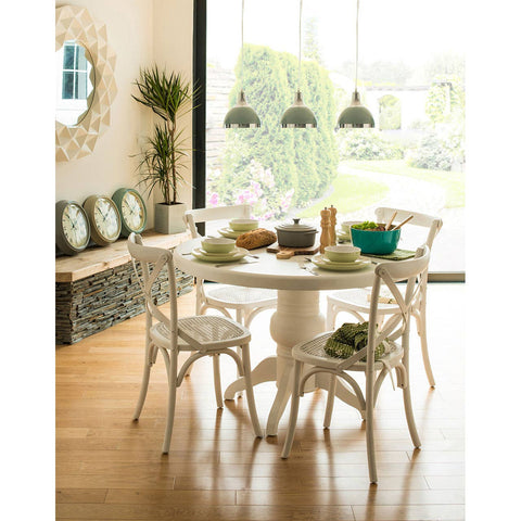 Round dining table with a white wash finish surrounded by four matching chairs, set in a bright room with large windows. The table is adorned with a decorative centerpiece, bowls, and green plants. A stylish mirror and decorative cushions are visible in the background, along with wooden flooring and pendant lights hanging above