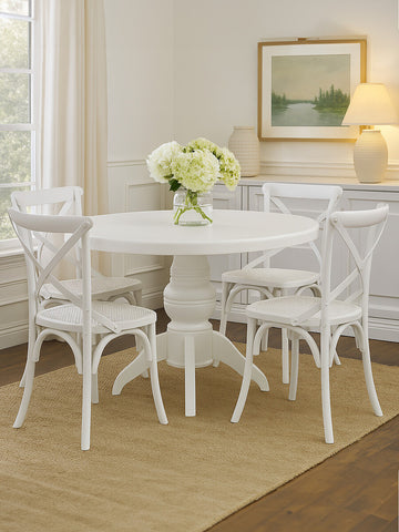 Round dining table with a white wash finish surrounded by four matching chairs, all featuring a classic design. A vase of white flowers sits on the table, and the setting includes a light-colored rug and a softly lit room with a framed landscape painting on the wall
