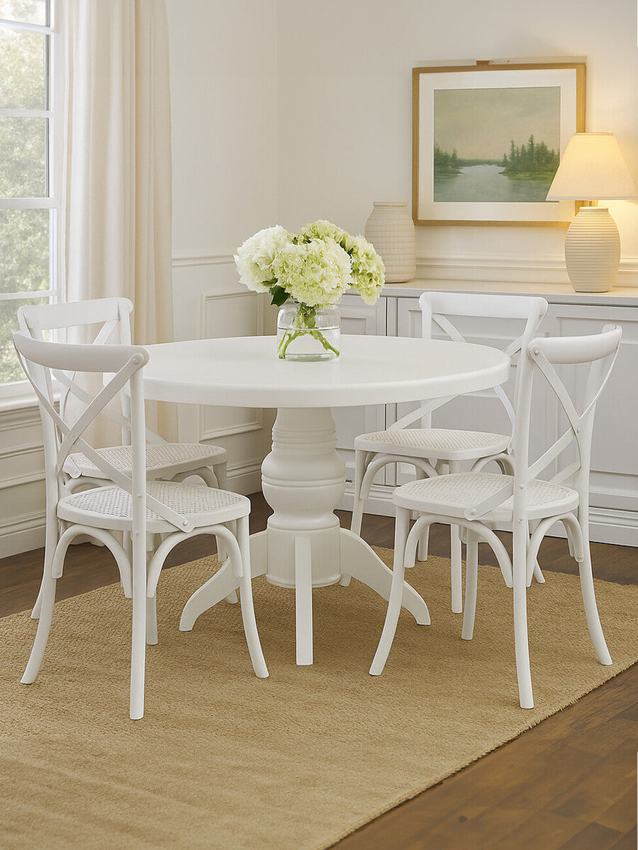 Round dining table with a white wash finish surrounded by four matching chairs, all featuring a classic design. A vase of white flowers sits on the table, and the setting includes a light-colored rug and a softly lit room with a framed landscape painting on the wall
