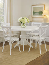 Round dining table with a white wash finish surrounded by four matching chairs, all featuring a classic design. A vase of white flowers sits on the table, and the setting includes a light-colored rug and a softly lit room with a framed landscape painting on the wall