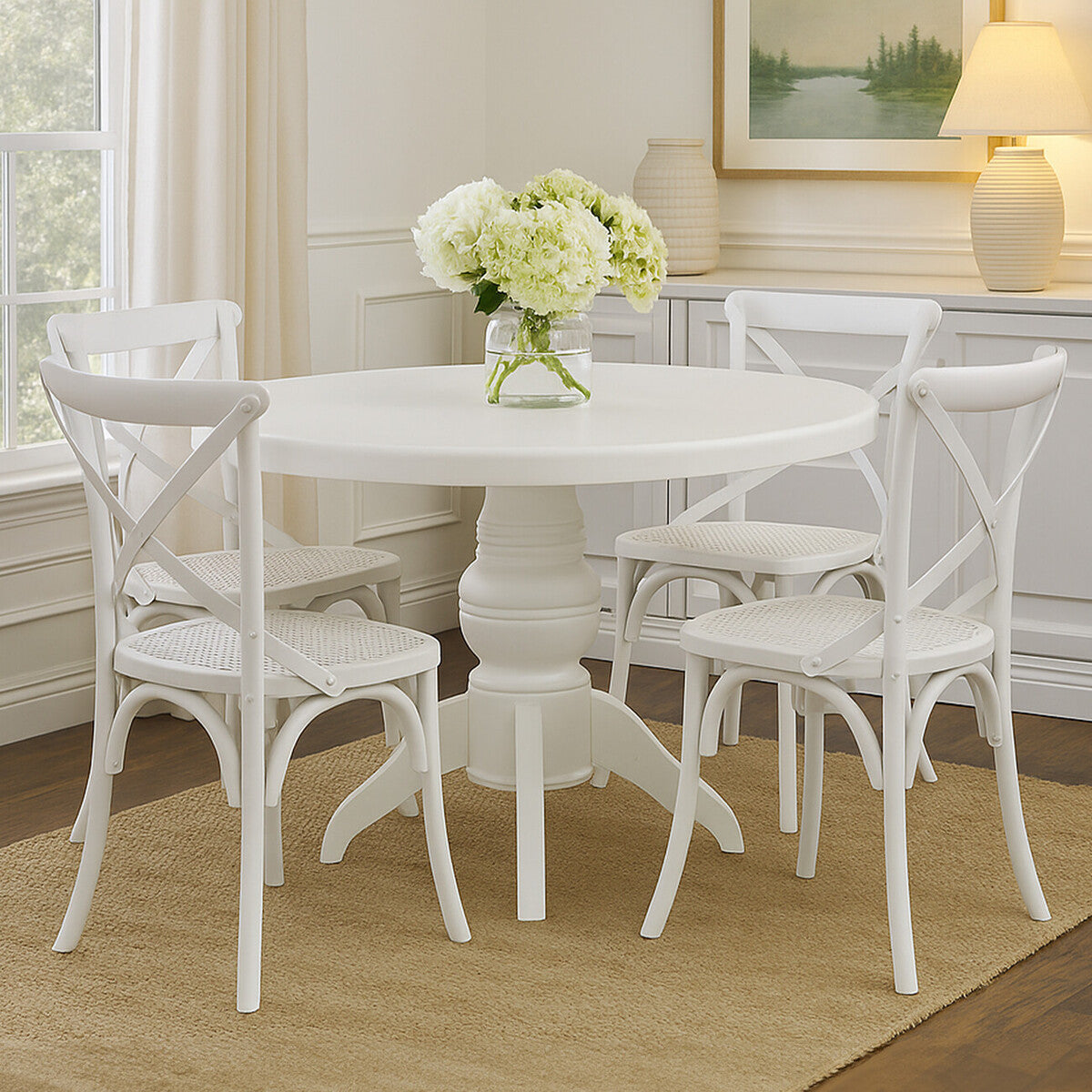 Round dining table with a white wash finish, accompanied by four matching chairs featuring a crisscross back design. A vase with white flowers sits at the center of the table. The setting includes a light-colored rug and large windows allowing natural light to fill the room