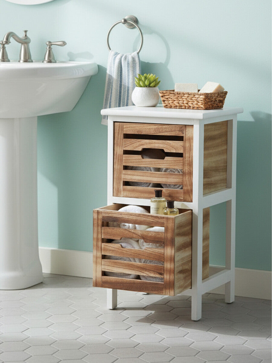 A white two-drawer chest with wooden slatted drawers, placed next to a sink in a light blue bathroom. The top of the chest features a small basket and a decorative item, while one drawer is partially open, revealing its interior. The floor is light-colored, complementing the overall aesthetic